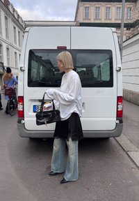 Persona con cabello rubio corto, camisa blanca holgada, falda negra de capas y jeans descoloridos, sosteniendo un bolso negro, de pie frente a una furgoneta blanca.