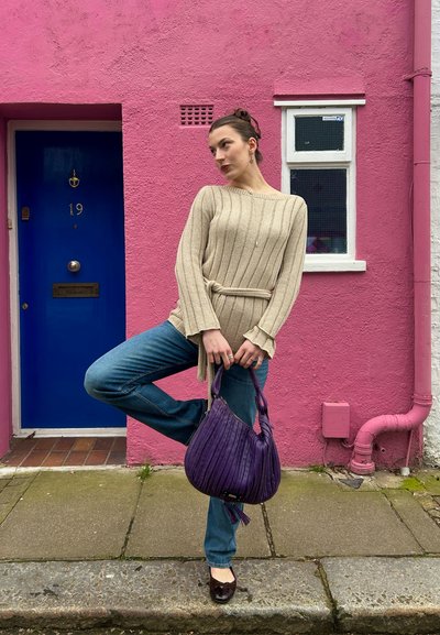 Mujer posa sobre una pierna frente a una pared rosa con una puerta azul, vistiendo un suéter beige, jeans azules, sosteniendo un bolso morado.