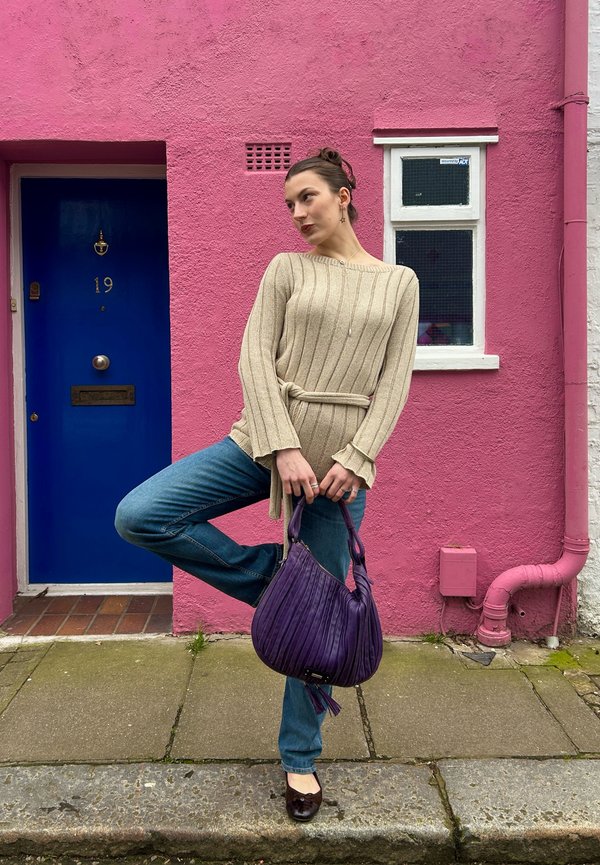 Donna posa su una gamba davanti a una parete rosa con una porta blu, indossa un maglione beige, jeans blu, e tiene una borsa viola.