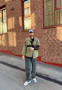 Ljusgrön jacka med svart armstöd över en svart skjorta, i kombination med mörkgröna cargo-byxor och grå sneakers, mot en tegelvägg.