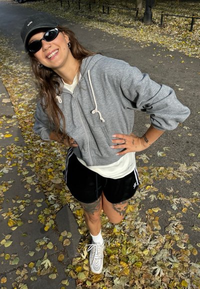 Sudadera gris con cremallera y cordones blancos, gorra negra, pantalones cortos negros con rayas blancas y zapatillas blancas en un camino cubierto de hojas de otoño.