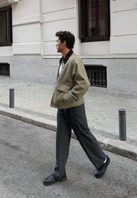 Veste oversize vert olive avec un col marron contrastant, associée à un pantalon plissé gris foncé et des chaussures noires, marchant dans une rue.