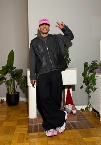 Veste en denim noir avec poches en cuir, pantalon noir ample et baskets blanches avec des touches de rose. Casquette rose et plantes en arrière-plan.