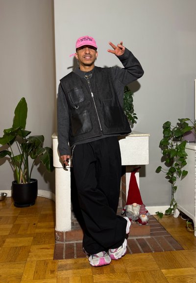 Chaleco de mezclilla negra con bolsillos de cuero, pantalones negros holgados y zapatillas blancas con detalles en rosa. Gorra rosa y plantas de fondo.