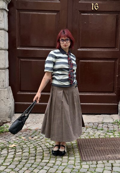 Camisa de rayas de manga corta con un cuello oscuro, combinada con una falda midi escocesa marrón. Un bolso negro y zapatos con detalles blancos completan el look.