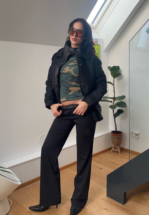 Kvinne iført stor svart jakke, camo crop top, svarte bukser, spisse sko, og store solbriller står innendørs ved siden av en potteplante.