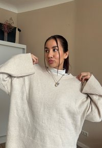 Pull oversized beige clair avec des poignets à côtes, présentant une texture douce. La personne porte un col roulé blanc en dessous et des colliers superposés.