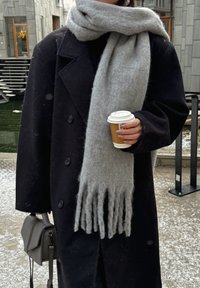 Casaco preto de lã com grandes botões, combinado com um lenço franjado cinza claro. Segurando um copo de café descartável e uma pequena bolsa cinza.