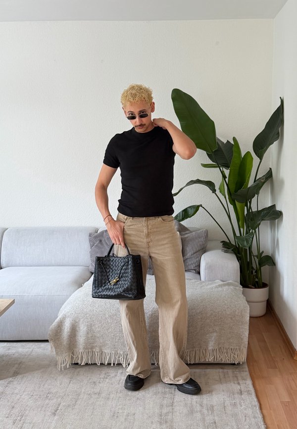 Mężczyzna o kręconych blond włosach, ubrany w czarny t-shirt i beżowe spodnie, trzyma czarną plecioną torebkę stojąc w salonie z sofą i dużą zieloną rośliną.