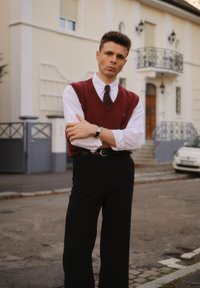 Mladý muž v bílém tričku, červené vestě a kravatě stojí s překříženýma rukama na městské ulici před béžovou budovou s balkonem.