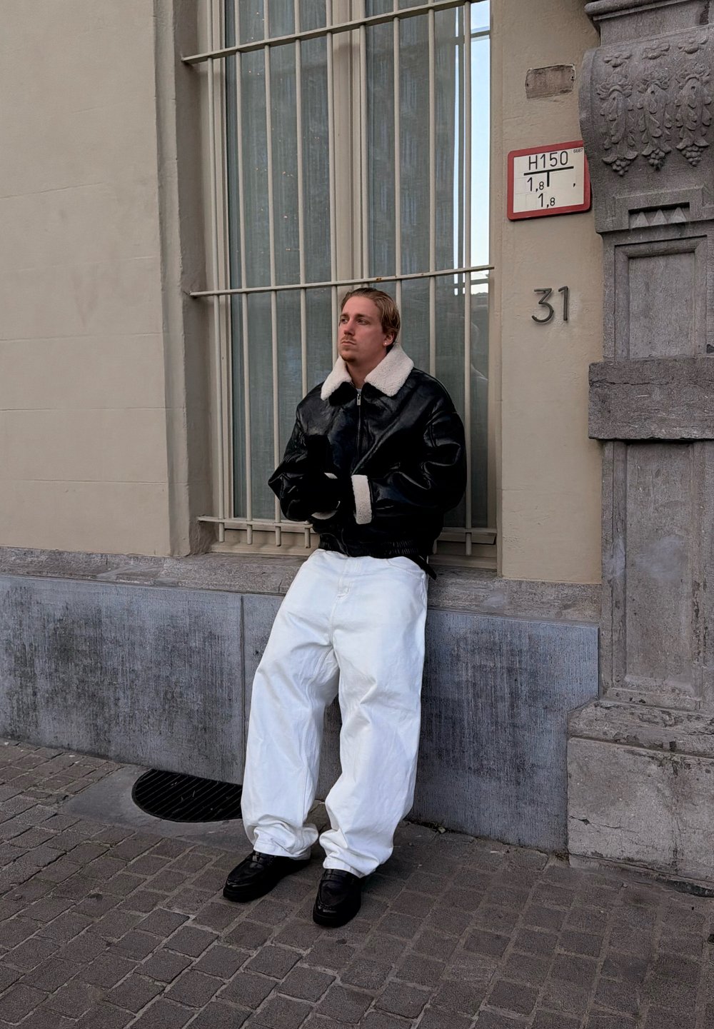 Homme appuyé contre une fenêtre avec barreaux dans une rue de la ville, portant une veste noire avec un col blanc, un pantalon blanc et des chaussures noires, près du bâtiment numéro 31.