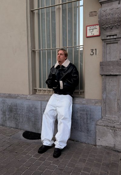 Hombre apoyado contra ventana con rejas en calle de la ciudad, vistiendo chaqueta negra con cuello blanco, pantalones blancos y zapatos negros, cerca del edificio número 31.