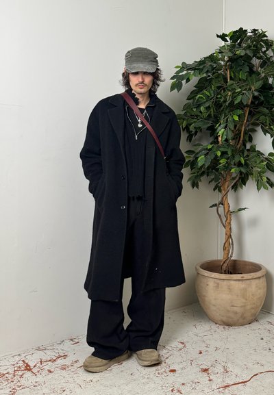 Hombre con abrigo negro de gran tamaño, pantalones negros, zapatos beige, gorra gris y bolso al hombro, de pie junto a una gran planta en maceta en el interior.