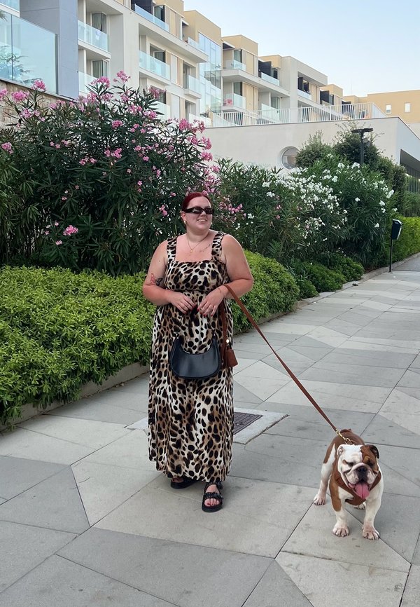 Robe maxi à imprimé léopard, sac à main noir, sandales sur un chemin pavé. Bulldog marron et blanc en laisse, entouré de verdure et de fleurs.