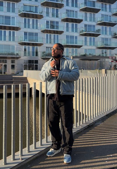 Hombre con gafas y cabello trenzado, vestido con una chaqueta clara y pantalones oscuros, de pie junto a una barandilla de metal cerca del agua, frente a un edificio moderno de cristal.