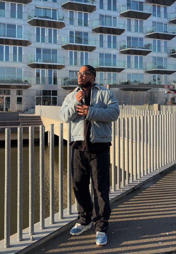 Homme portant des lunettes et les cheveux tressés, vêtu d'une veste légère et d'un pantalon foncé, se tient près d'une rambarde en métal près de l'eau devant un bâtiment moderne en verre.