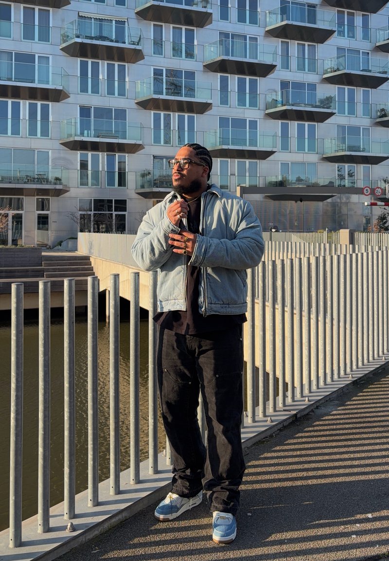Homme portant des lunettes et les cheveux tressés, vêtu d'une veste légère et d'un pantalon foncé, se tient près d'une rambarde en métal près de l'eau devant un bâtiment moderne en verre.