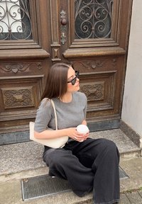 Top de punto gris de manga corta, pantalones negros anchos, sosteniendo una taza de café blanca. Bolso cruzado de color claro. Sentada cerca de una puerta de madera ornamentada.
