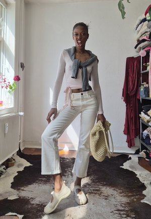 Mujer posando en interiores con pantalones blancos, una camiseta de manga larga rosa claro, un suéter gris atado alrededor de su cuello y sosteniendo un bolso beige tejido.