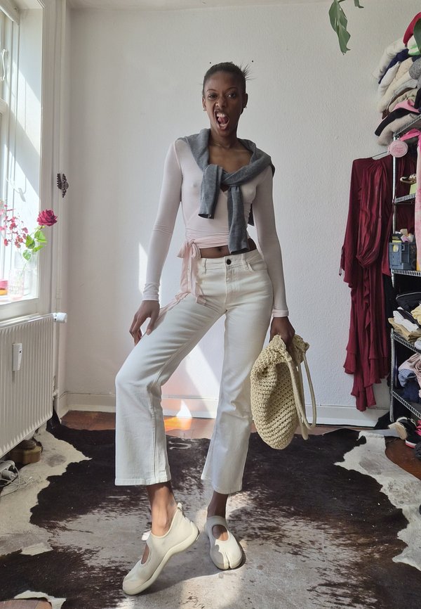 Vrouw poseert binnen in een witte broek, een lichtroze top met lange mouwen, een grijze trui om haar nek gebonden en houdt een beige geweven tas vast.