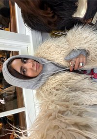 Femme portant une capuche en maille grise et un manteau crème duveteux, tenant un téléphone, avec deux bagues argentées et du vernis à ongles rouge sur sa main.