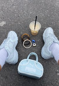 Fehér New Balance sportcipők pöttyös zoknikkal, jeges kávé fekete szívószállal, karkötők, gyűrűk, és egy világoskék Puma táska az aszfalton.