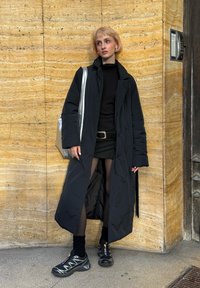 Svart puffjakke med lang design og vide ermer, brukt over en svart topp og skjørt. Sølvfarget totebag og svarte joggesko fullfører antrekket.
