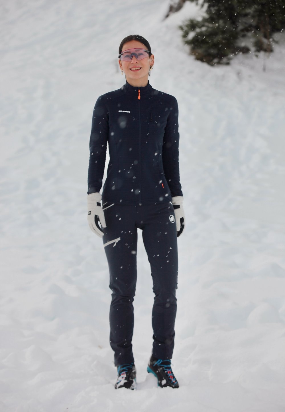 Combinaison de ski intégrale bleue avec une fermeture éclair, des accents orange et des gants. Sol couvert de neige avec des flocons de neige tombants en arrière-plan.