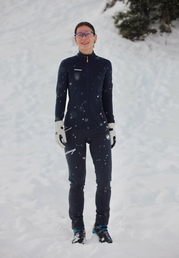 Tuta da sci blu a corpo intero con zip, dettagli arancioni e guanti. Terreno coperto di neve con fiocchi di neve che cadono sullo sfondo.