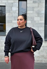 Czarny sweatshirt Nike, długie rękawy, luźny krój. W połączeniu z burgundową spódnicą, trzymając burgundową torbę. Gładkie tekstury, minimalne logo.