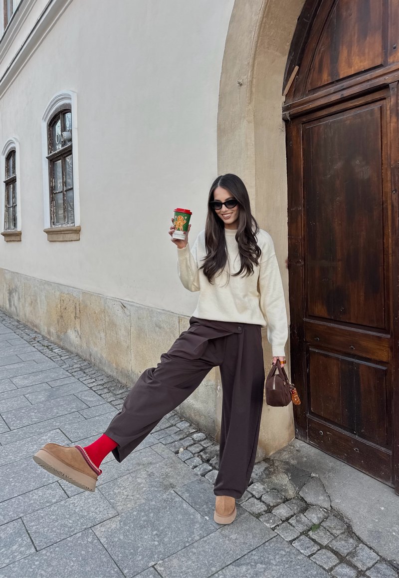 Femme aux longs cheveux noirs, portant des lunettes de soleil, un pull crème et un pantalon large marron, tenant une tasse festive et un petit sac à main marron près d'une porte en bois.