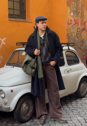Un joven con un abrigo azul marino, bufanda verde y suéter estampado está parado junto a un coche vintage blanco contra una pared naranja.