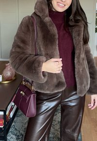 Femme portant une veste en fausse fourrure marron, un pull à col roulé bordeaux, un pantalon en cuir marron foncé, et portant un sac à main bordeaux.