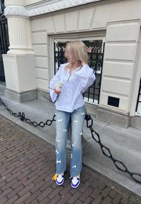 Camisa blanca oversized con mangas acampanadas, jeans color azul claro adornados con lazos de tela y zapatillas moradas y blancas. Sosteniendo una taza.