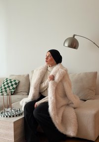 Abrigo de piel sintética en beige claro, suéter de punto acanalado en crema, pantalones negros con rayas verticales, sentado en un sofá texturizado con un cojín a cuadros verde.