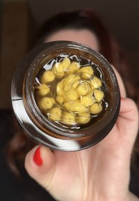 Brunt glasburk fylld med gula kapslar i klart vätska, rund form, metallisk reflekterande kant, hålls i hand med rödfärgat nagellack.