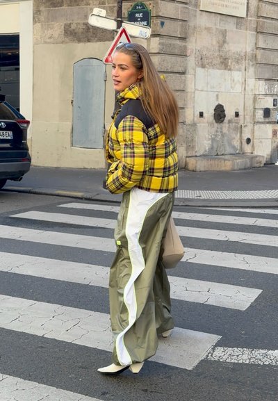 Mujer de cabello largo que lleva una chaqueta acolchada amarilla a cuadros y pantalones verdes anchos cruzando una calle de la ciudad por un paso de peatones marcado.