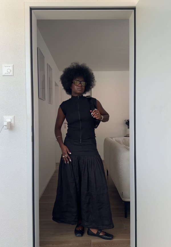 Femme aux grands cheveux bouclés portant un haut noir sans manches, une longue jupe noire, des lunettes et des chaussures noires, debout dans un encadrement de porte à l'intérieur d'une pièce moderne.
