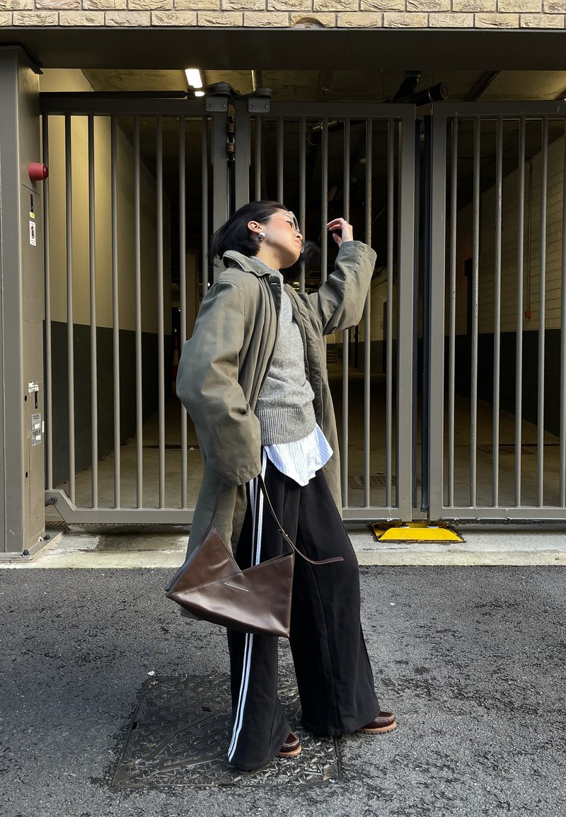 Personne portant une veste verte oversize, un pull gris, un pantalon large noir avec des rayures blanches, tenant un sac triangulaire marron, debout près d'une grille en métal sur l'asphalte.