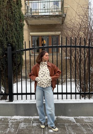 Mujer con chaqueta marrón, suéter de lunares, vaqueros y zapatillas de deporte de pie en una acera nevada frente a una cerca de metal negra y un edificio beige.