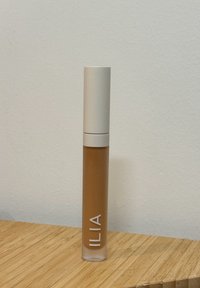 Produit liquide ILIA dans un tube transparent avec un capuchon beige clair. Le liquide est d'une teinte marron chaud avec une texture lisse.