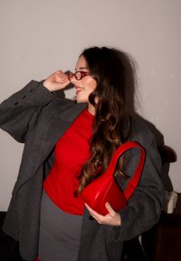 Femme aux longs cheveux bruns portant des lunettes rouges, un haut rouge, une jupe grise et un manteau gris surdimensionné, tenant un sac à main rouge et souriant sur le côté.
