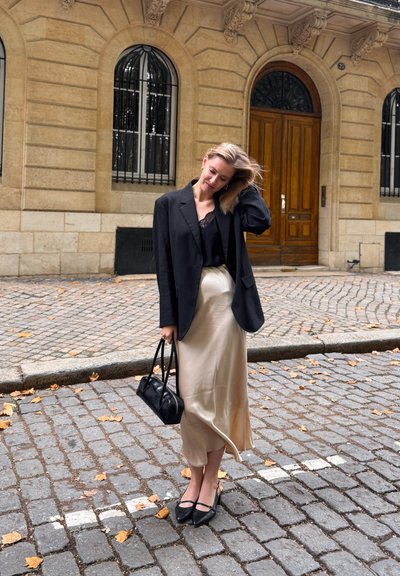 Blazer negro sobre una blusa de encaje negra, combinado con una falda midi beige de satén, bolso negro y zapatos negros en una calle de adoquines.