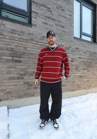 Hombre con camisa roja de manga larga a rayas, pantalones negros y zapatillas de deporte, de pie sobre la nieve frente a un edificio de ladrillos gris.