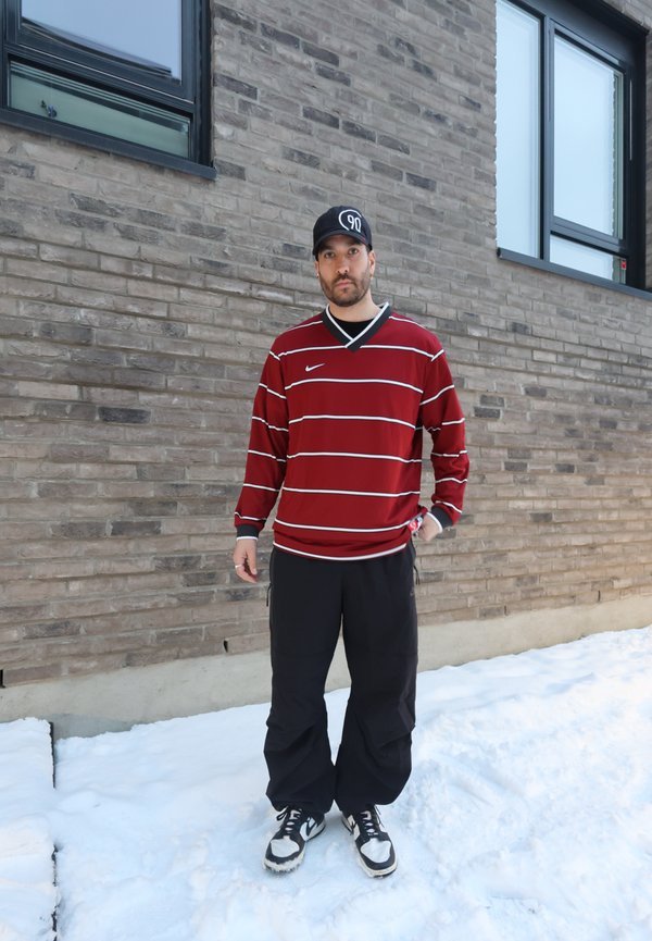 Uomo che indossa una maglia a maniche lunghe rossa a righe, pantaloni neri e scarpe da ginnastica, in piedi sulla neve davanti a un edificio in mattoni grigi.