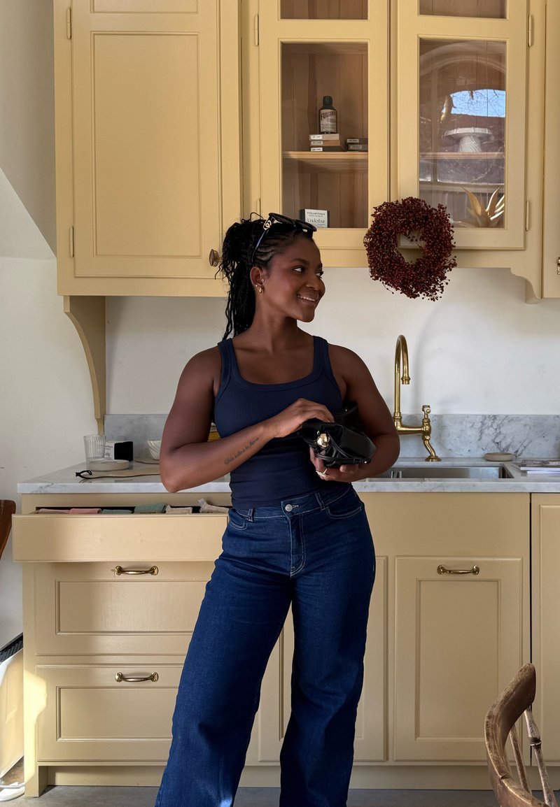 Femme aux cheveux tressés et lunettes de soleil sur la tête debout dans une cuisine jaune tenant un sac noir près d'un évier avec un robinet en laiton et une couronne rouge.