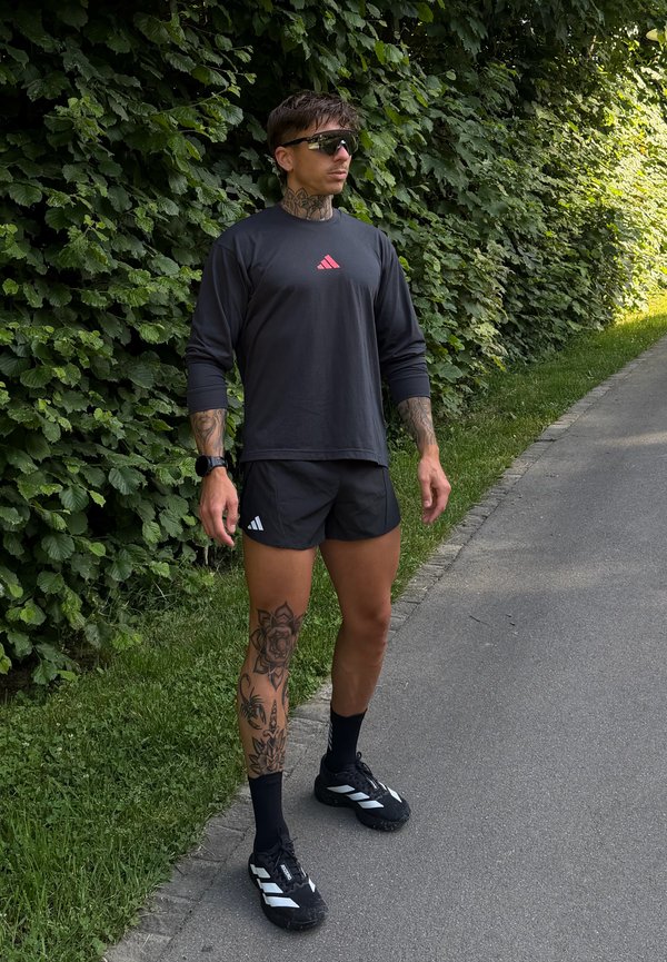 Mann med tatoveringer står på en sti ved grønne busker, med solbriller, svart Adidas-skjorte og shorts, svarte sokker og joggesko.