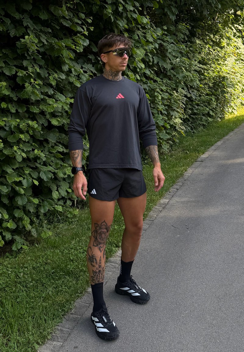 Homme avec des tatouages debout sur un chemin à côté de buissons verts, portant des lunettes de soleil, un t-shirt et un short noirs Adidas, des chaussettes noires et des chaussures de course.