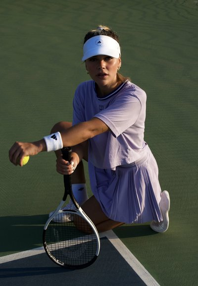 Jugadora de tenis vestida con un conjunto deportivo de lavanda, con falda de pliegues y top de manga corta, sosteniendo una raqueta y preparándose para sacar una pelota de tenis amarilla.