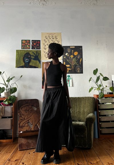 Top negro sin mangas con un diseño ajustado, combinado con una falda larga negra. De pie junto a una silla verde tapizada en una habitación con plantas y arte.